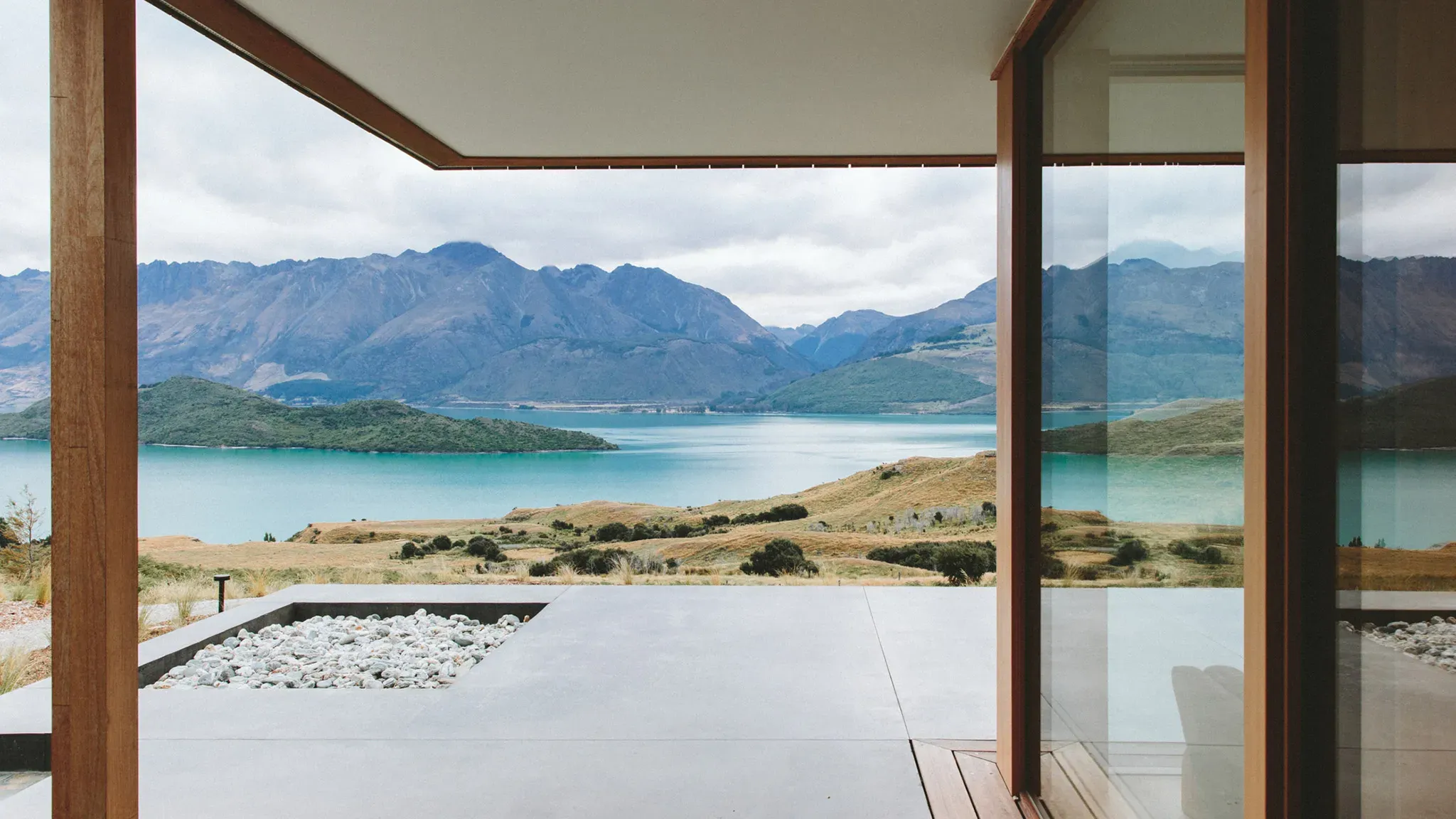 View of a lake surrounded by hills and mountains from a modern building with wooden frames and concrete flooring.