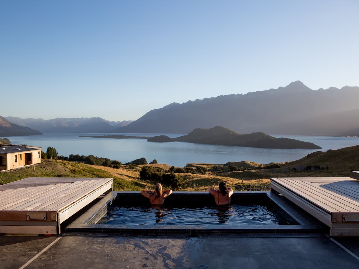 Two guests relaxing in a hot tub overlooking a lake and distant mountains at Aro Ha.