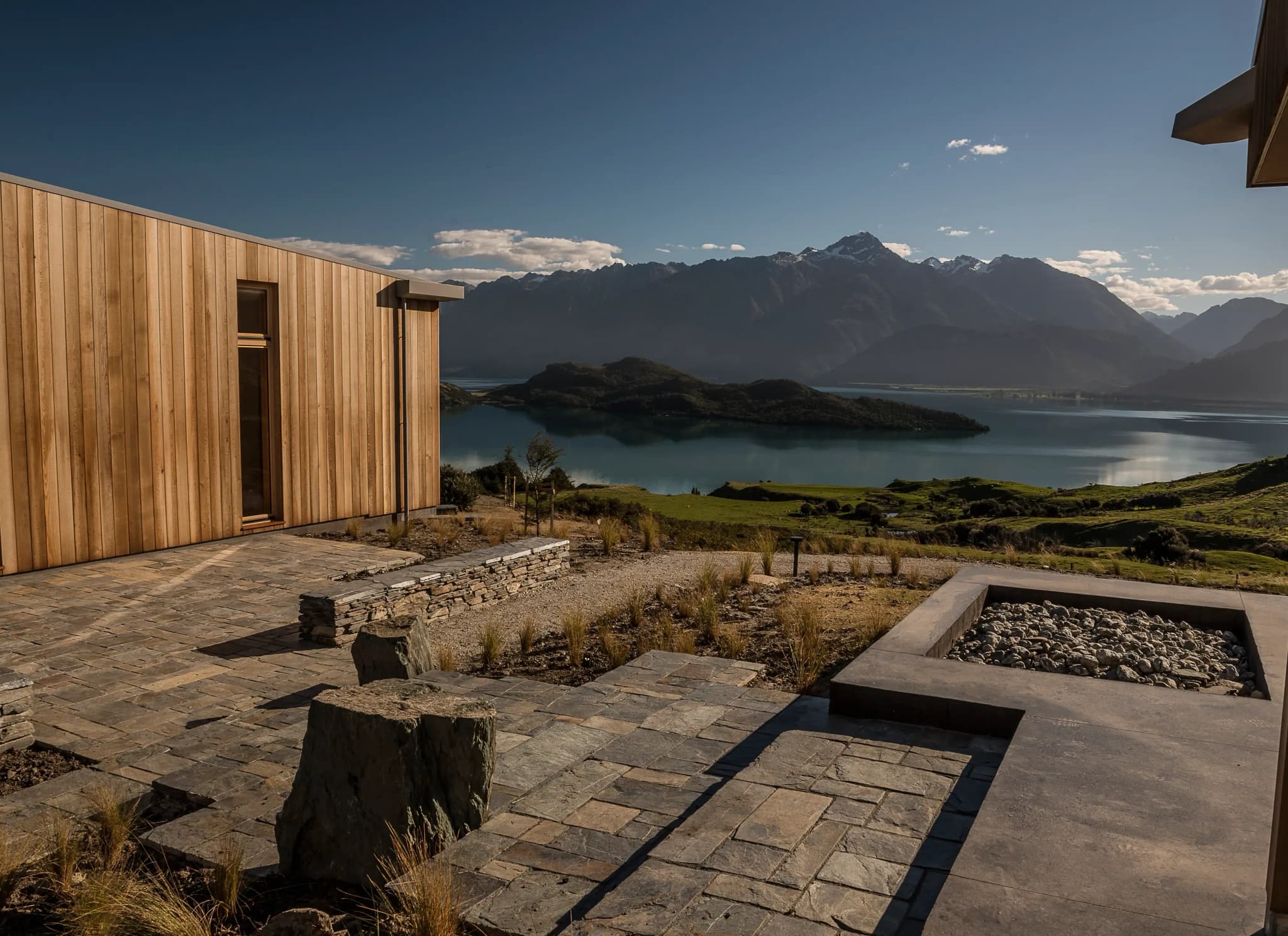 Timber-clad building exterior with stone pathways, rocky seating areas and views of the lake and mountains.