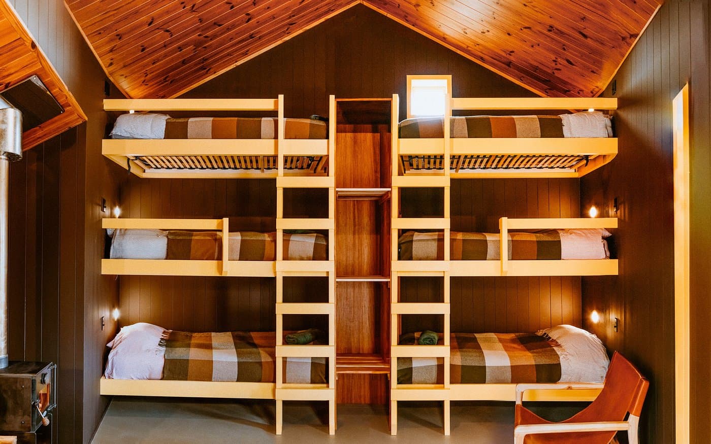 Symmetrical bunk room with six made beds, yellow ladders, timber ceiling and soft wall lighting.