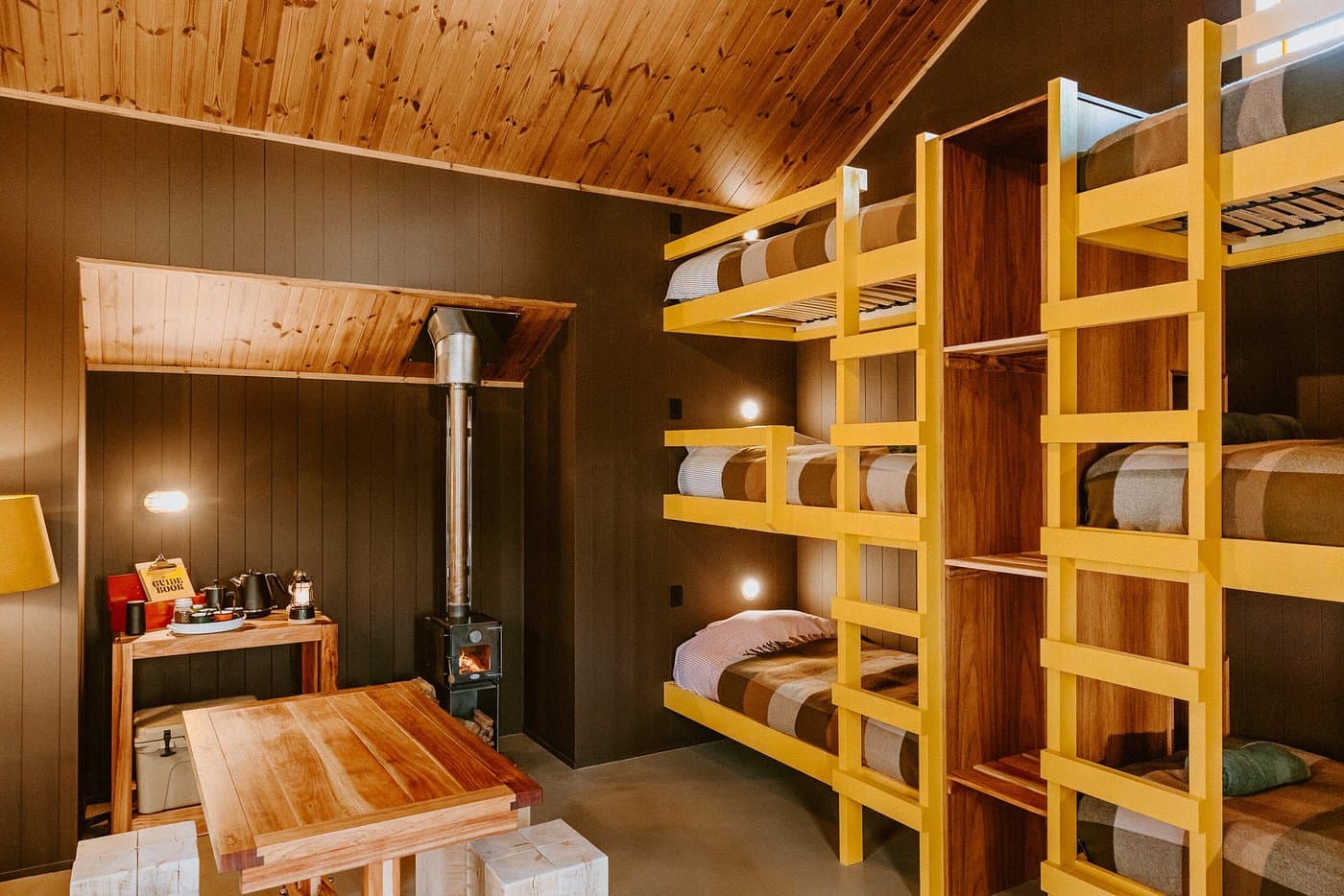 Warm timber bunk hut with yellow ladders, wood-burning fire, dining table and layered bedding.