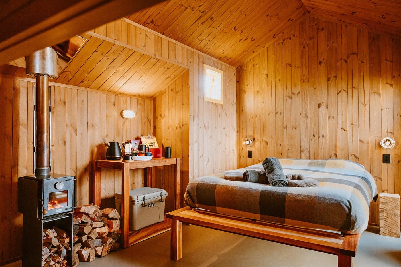 Cosy timber hut bedroom with a made bed, wood-burning fire, leather chair and warm alpine styling.