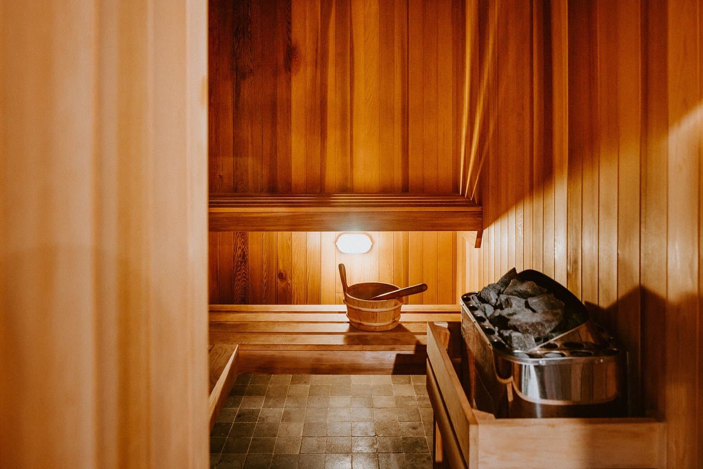 Timber sauna interior with wooden bench seating, sauna stones and bucket under warm ambient light.