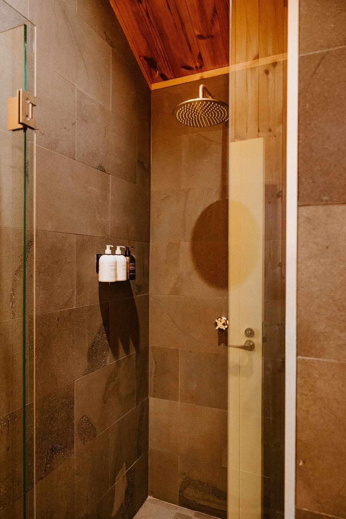 Private shower with stone tiles, timber ceiling, glass door and rainfall shower head.