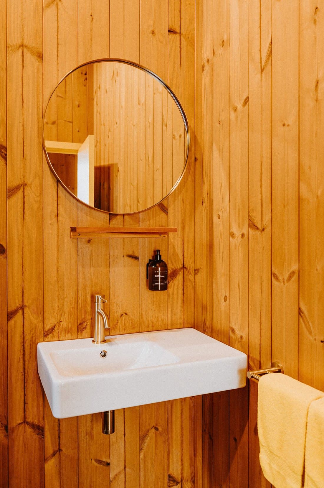 Timber bathroom vanity with round mirror, white basin, wall-mounted soap and towel rail.