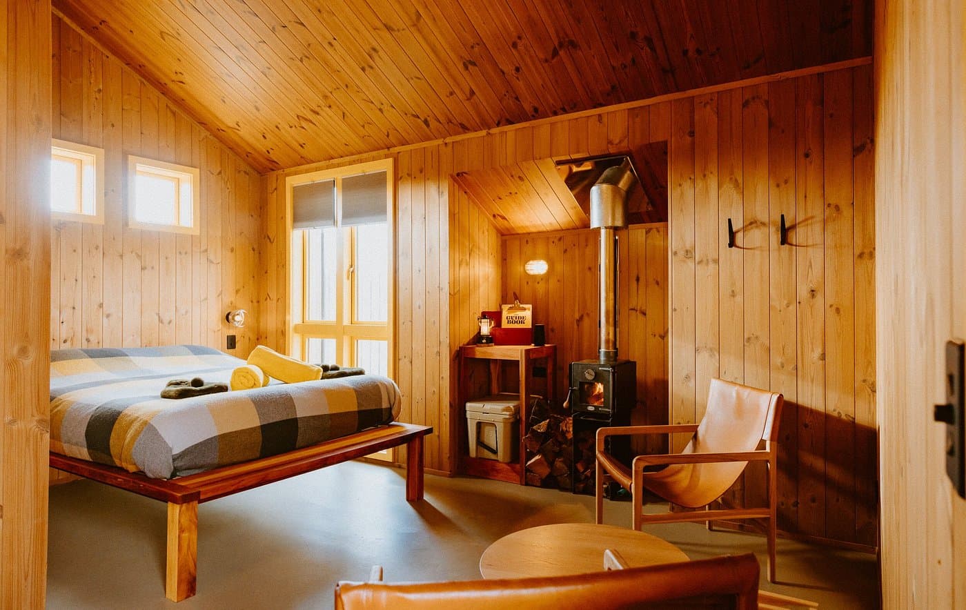 Self-contained timber hut with a made bed, wood-burning fire, seating area and warm natural finishes.