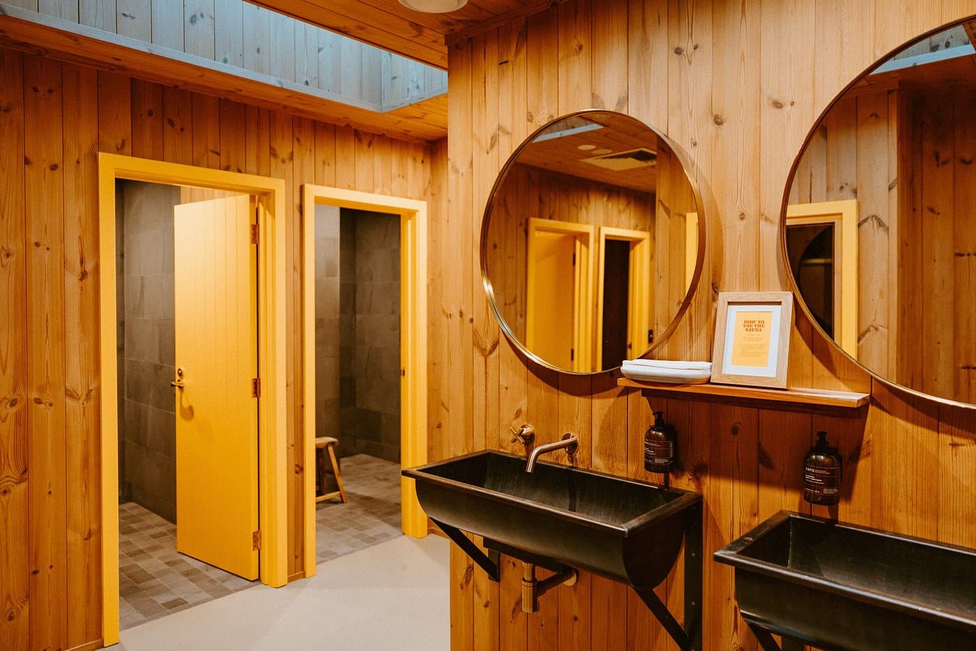 Shared bathroom with timber walls, black basins, round mirrors and yellow-trimmed shower doors.