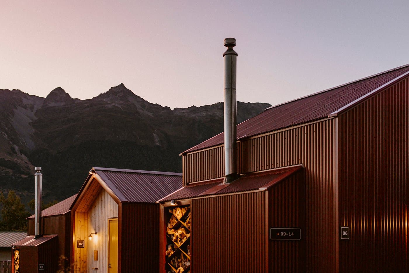 Alpine timber huts at The Great Glenorchy Alpine Base Camp, with wood stores, chimneys and mountain peaks at dusk.