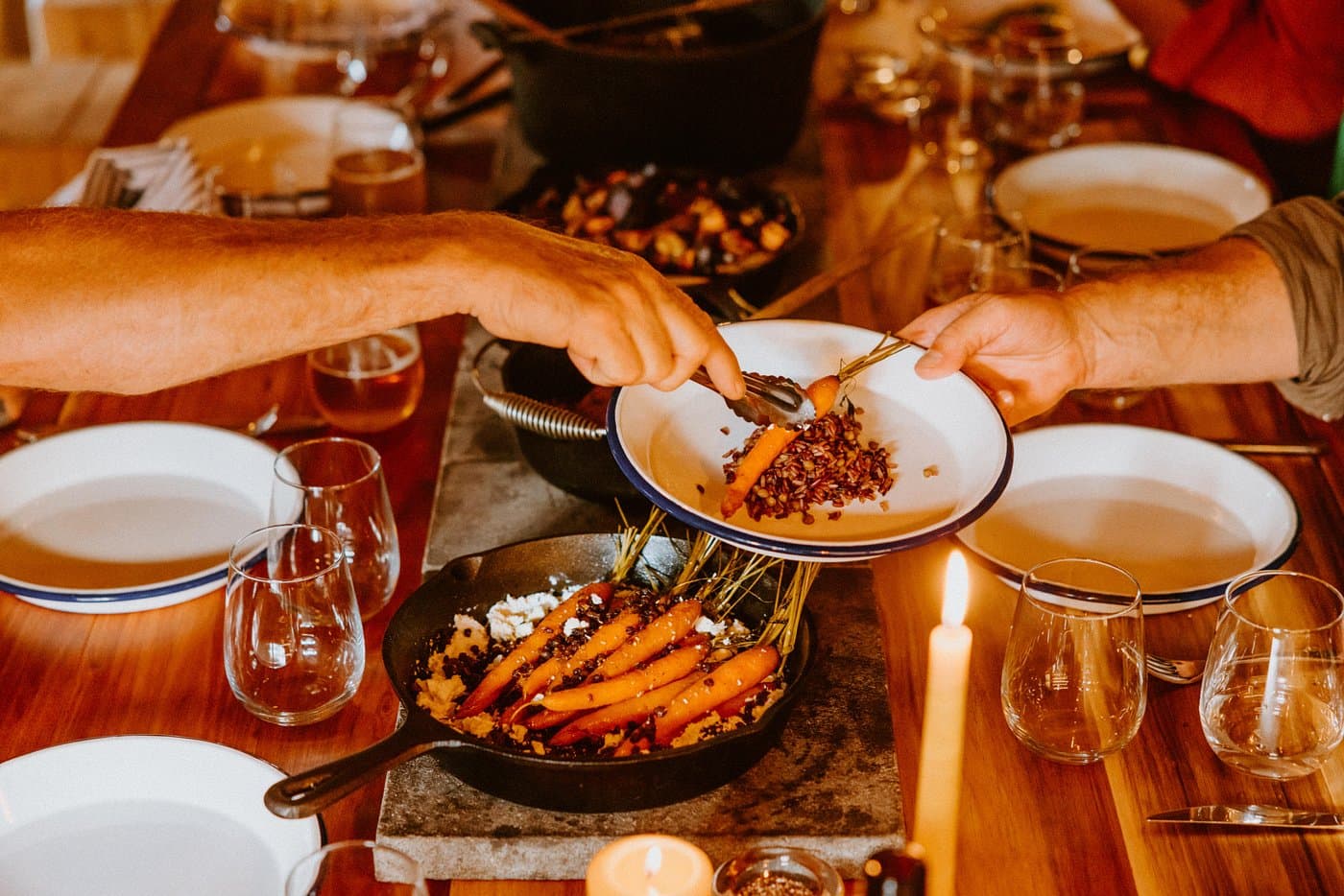 A guest serving roasted carrots and grains from cast-iron cookware at a candlelit dining table.
