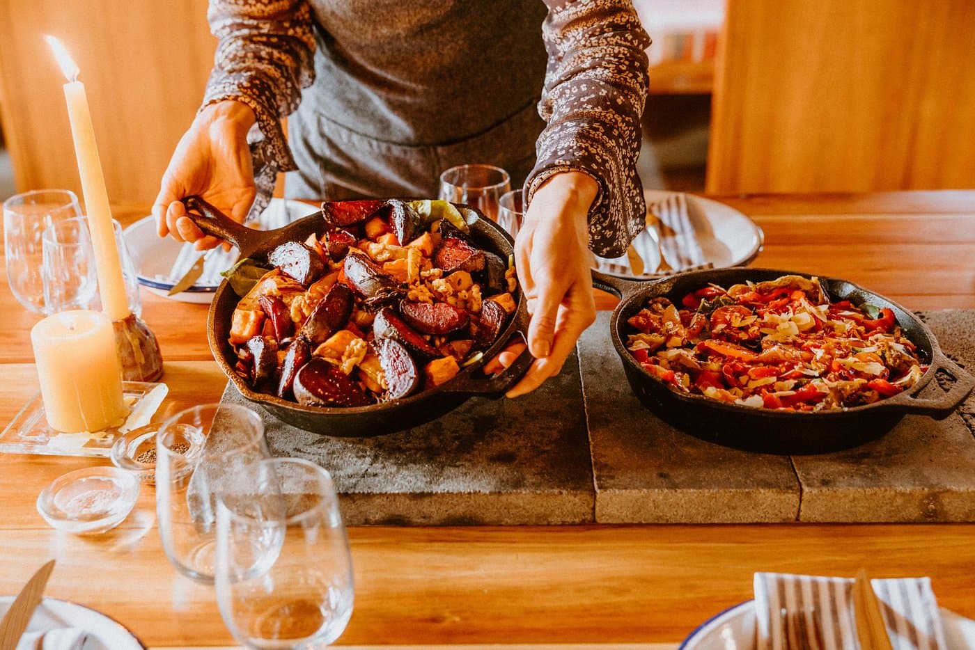 Cast-iron dishes of roasted vegetables being served at a warm timber dining table.