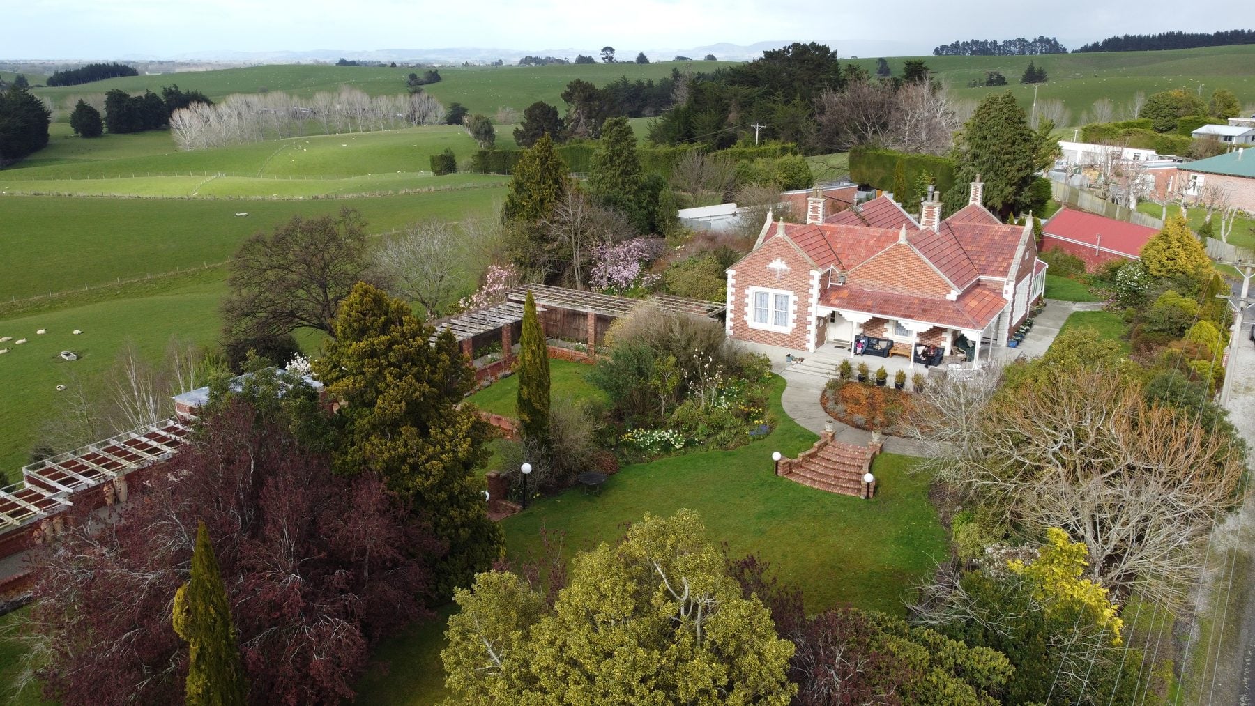 Heritage villa surrounded by gardens, featuring brick exterior, manicured lawns, and trees, set against rolling green hills.