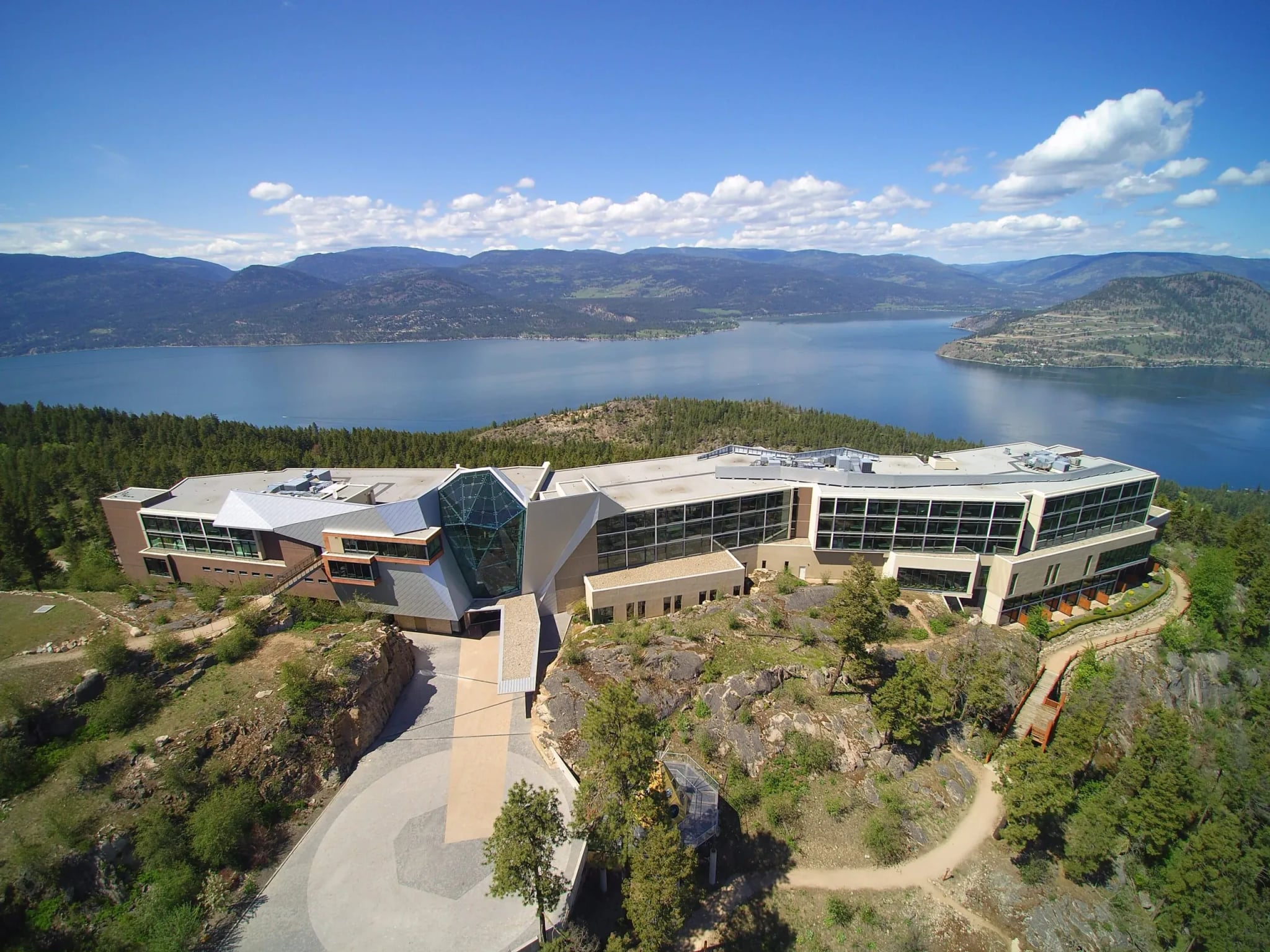 Aerial view of Sparkling Hill Resort with glass and stone architecture, surrounded by trees and overlooking Okanagan Lake.