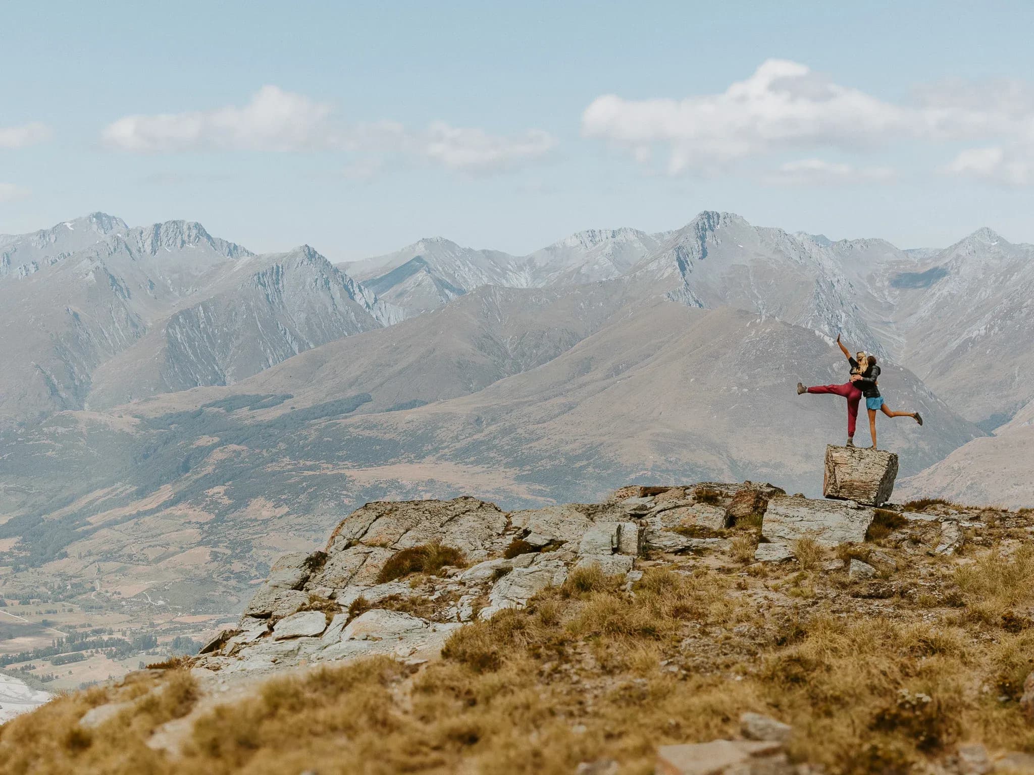 Two guests playfully pose on a rock outcrop with mountain ranges in the background under a blue sky.
