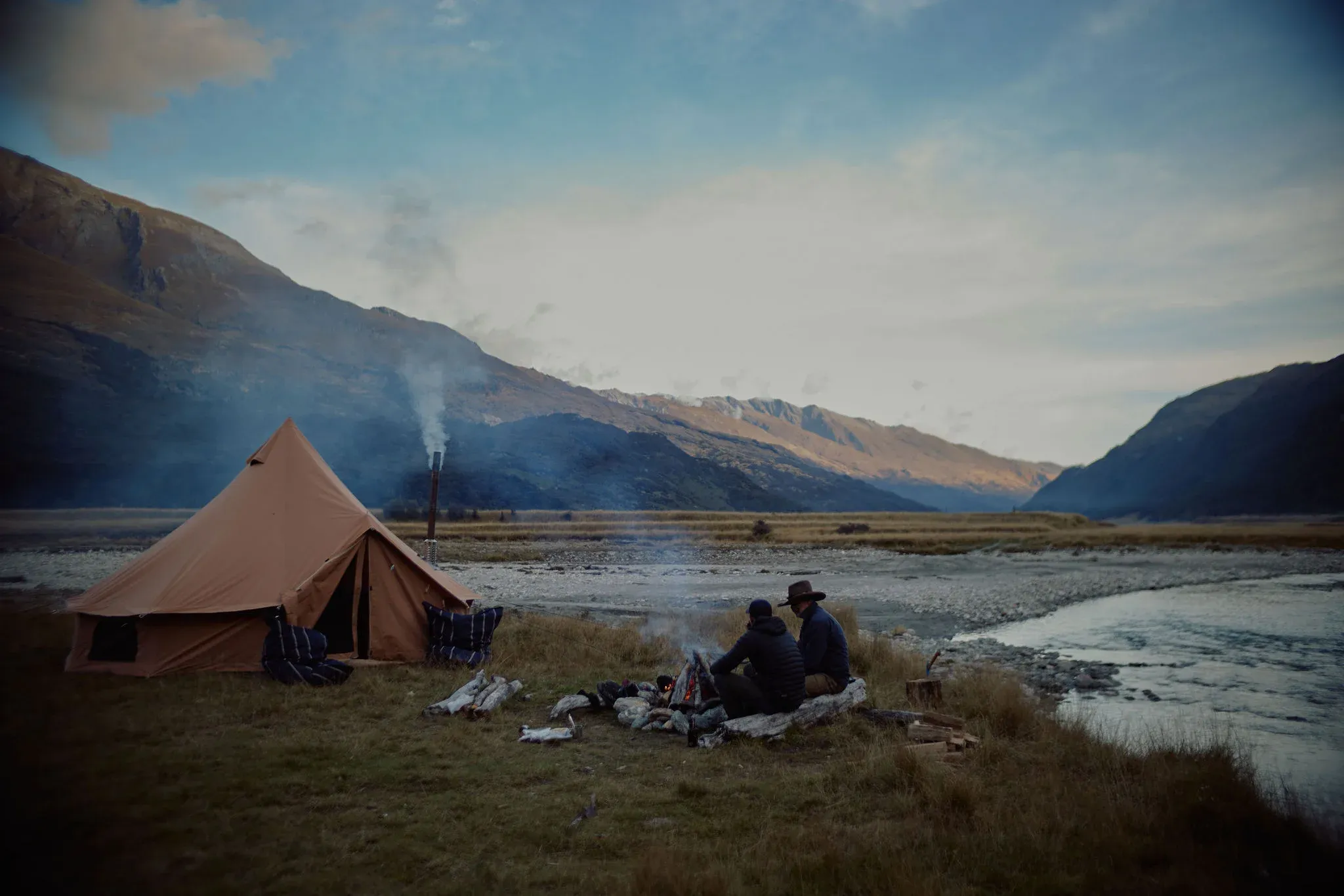 Tent set up near a river, with two guests sitting by a fire under a mountain backdrop.
