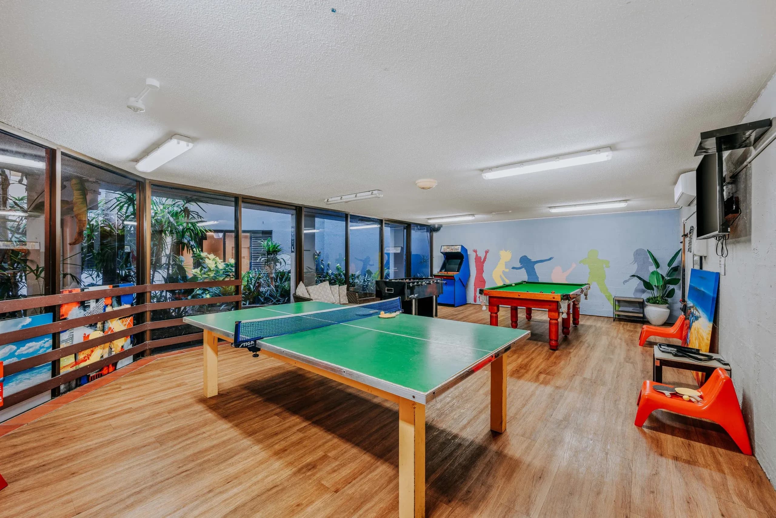 Recreation room with green ping pong table, red pool table, arcade machine, and large windows overlooking greenery.