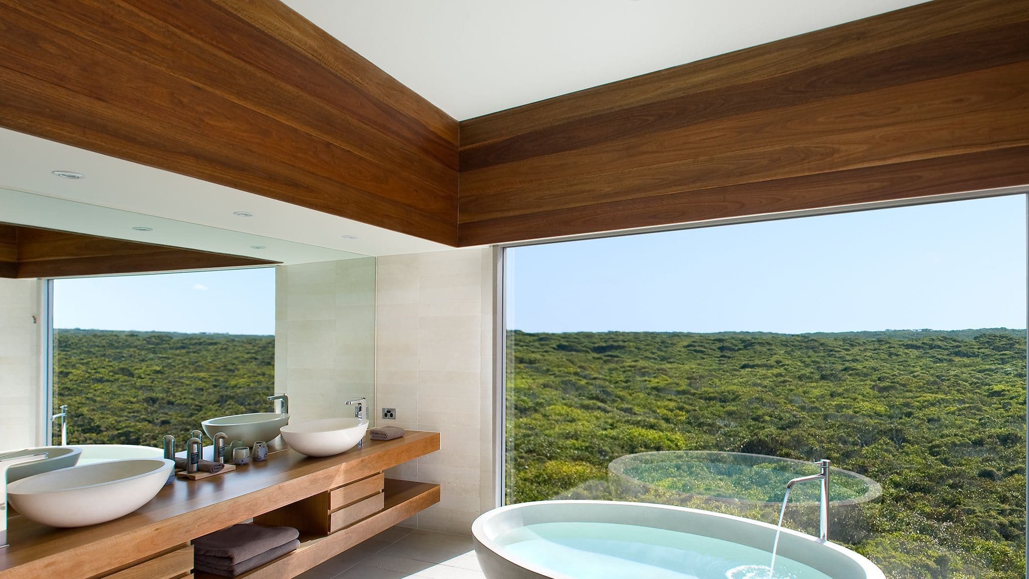 Bathroom interior featuring a freestanding round bathtub, wooden vanity with double sinks, and a large window overlooking greenery.