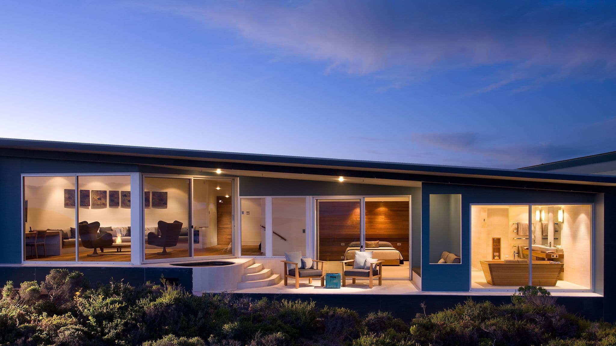 Lodge exterior at dusk, featuring large windows, a terrace with seating, and landscaped vegetation.