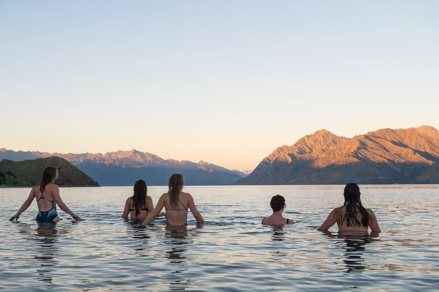 Guests taking a cold-water dip in Lake Hāwea, with mountain views at sunset.