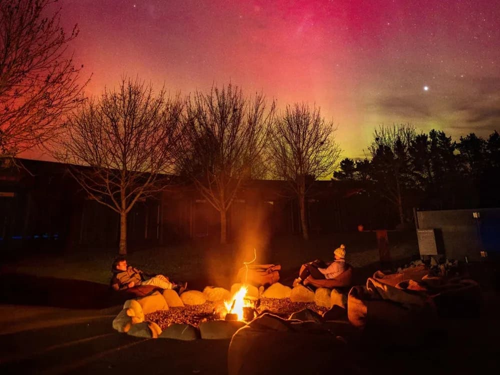 Guests seated around a fire pit with stones, under a colorful aurora night sky at Manawa Retreat.