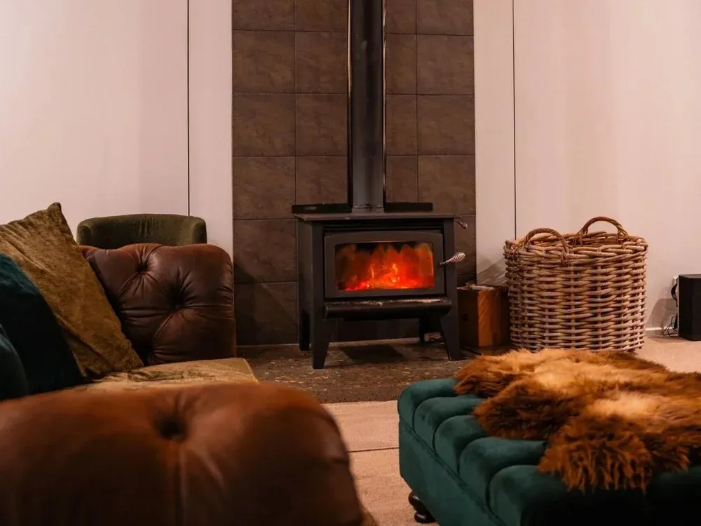 Cozy seating area featuring a green velvet ottoman, brown leather sofa, and a wood-burning stove against a tiled wall.