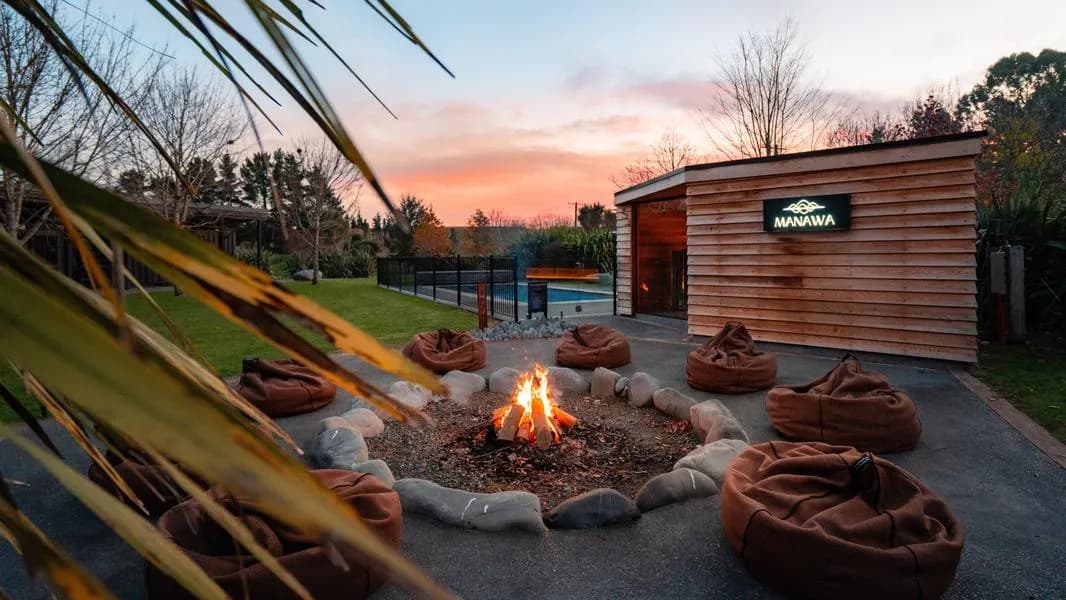 Fire pit surrounded by bean bags and stone seating, with a wooden building and sunset sky in the background.