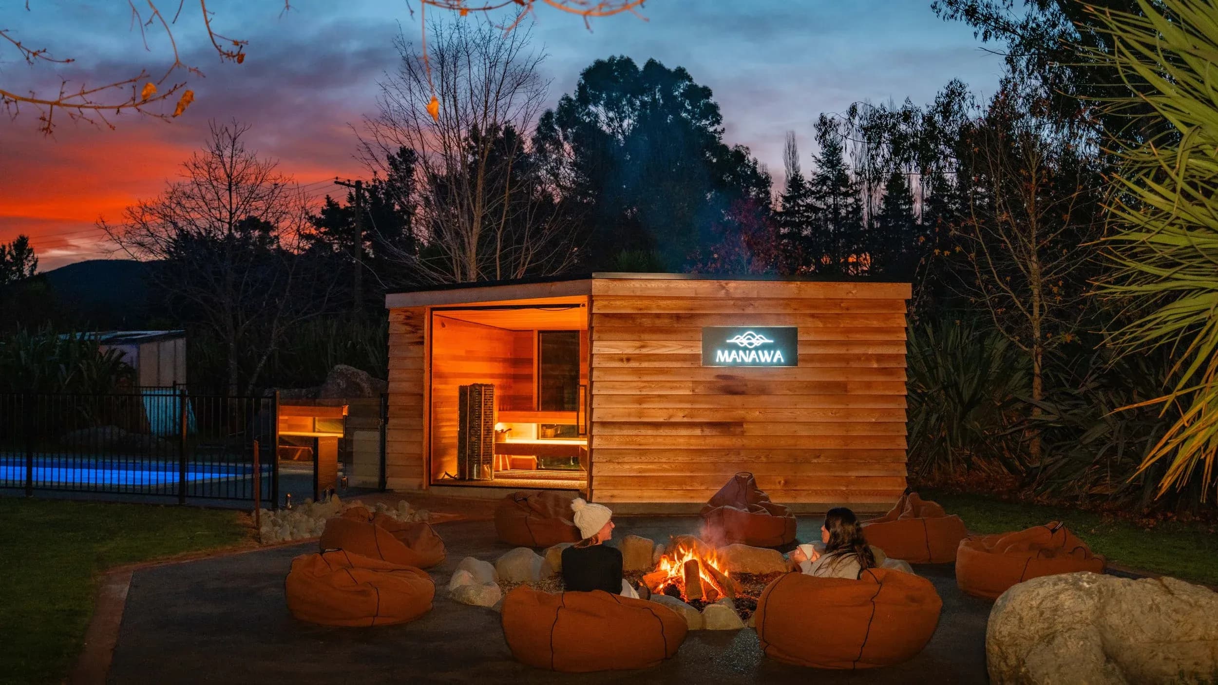 Guests sitting on bean bags around a fire pit in front of the sauna cabin at sunset.