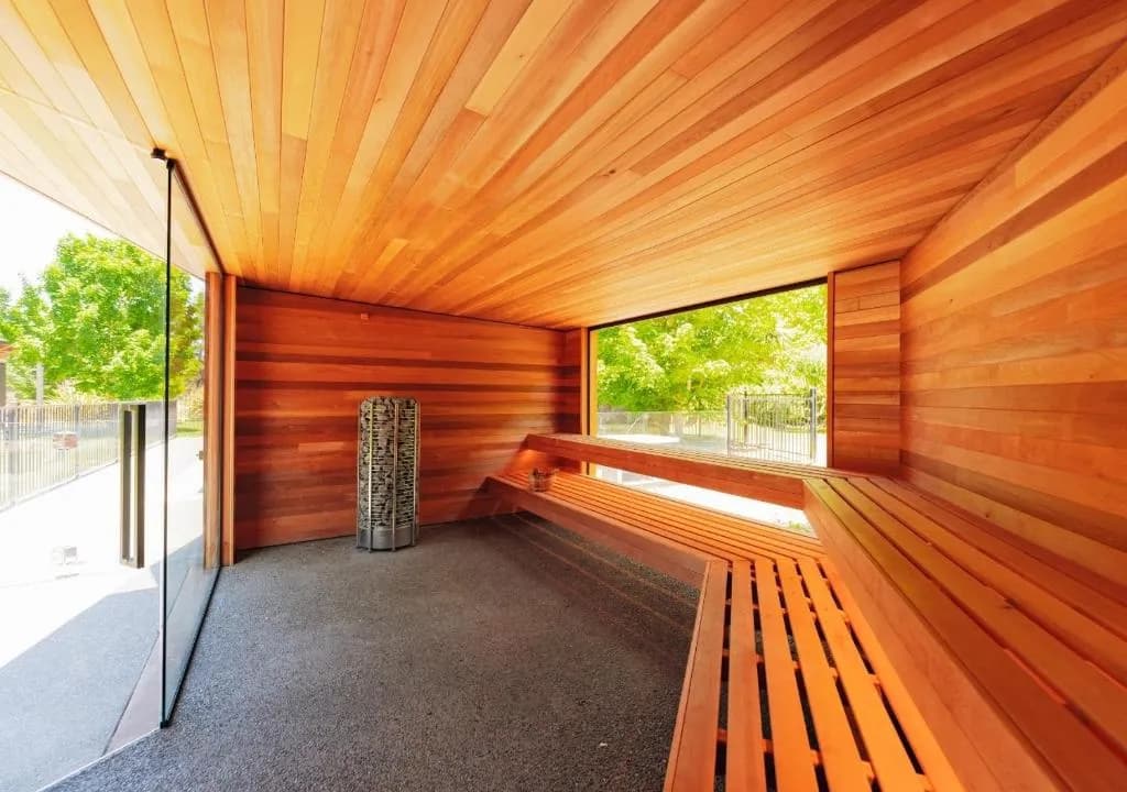 Timber sauna interior with wooden benches, stone heater, and large glass windows overlooking greenery.