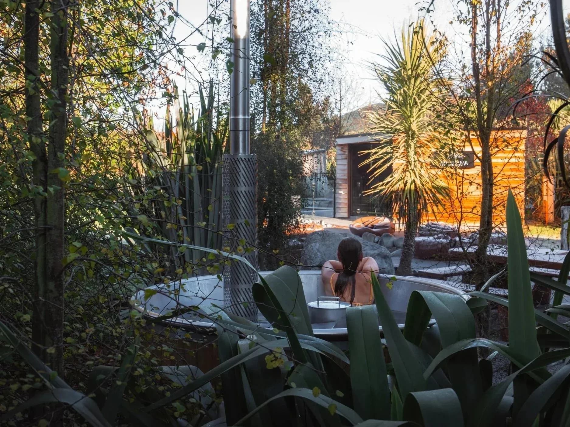 A guest relaxing in a hot tub surrounded by greenery and trees, with a wooden building in the background.