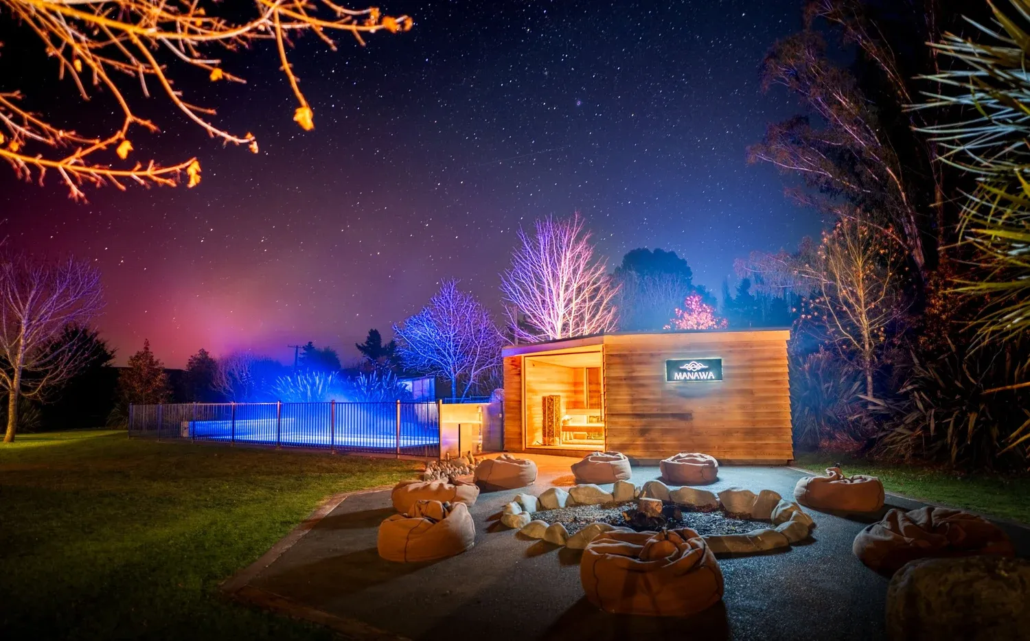 Manawa Retreat exterior at night, featuring a fire pit with seating, illuminated trees, and a starry sky with the aurora australis visible.