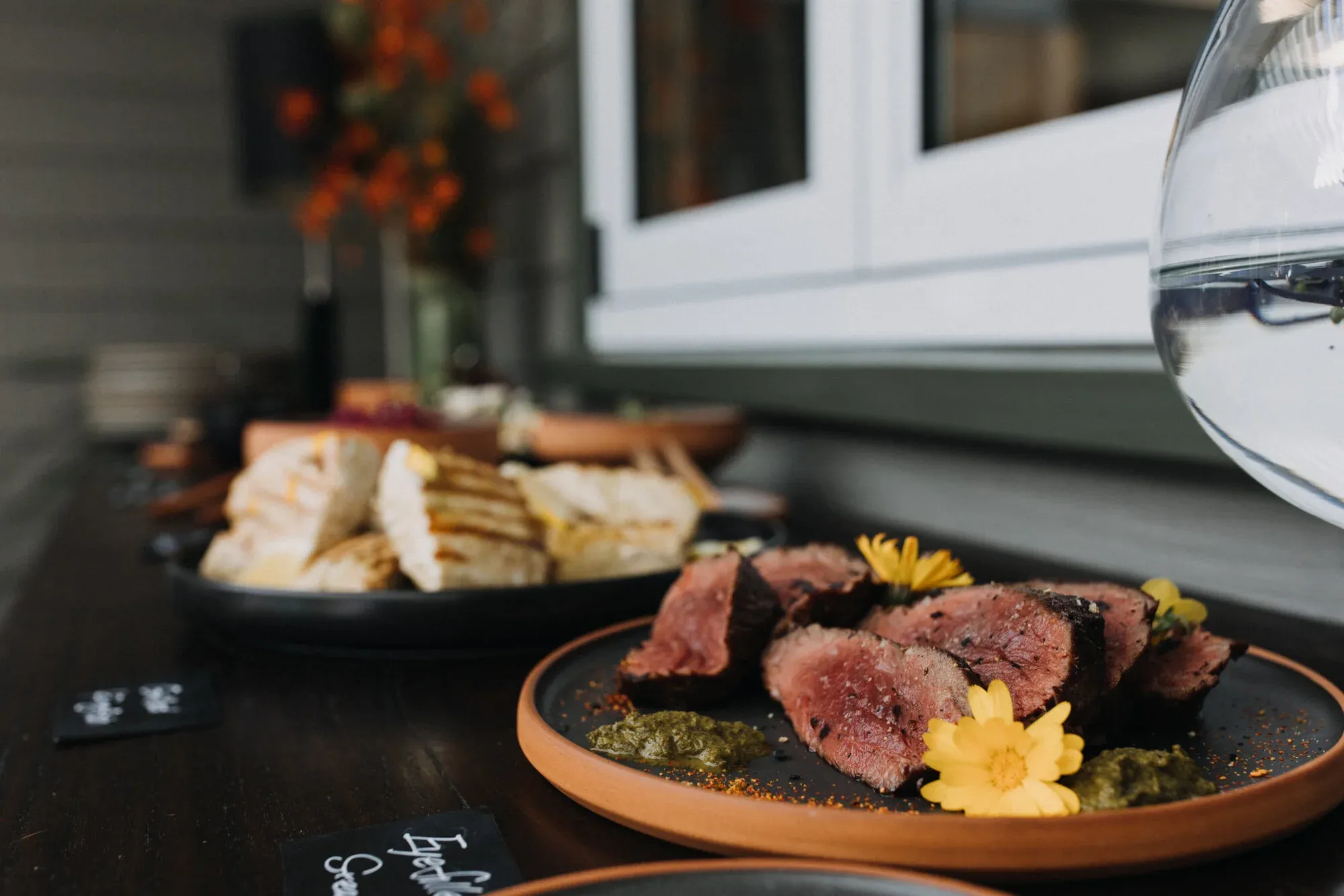 Plates of sliced meat garnished with flowers, accompanied by bread and small bowls of sauce on a dark wooden table.
