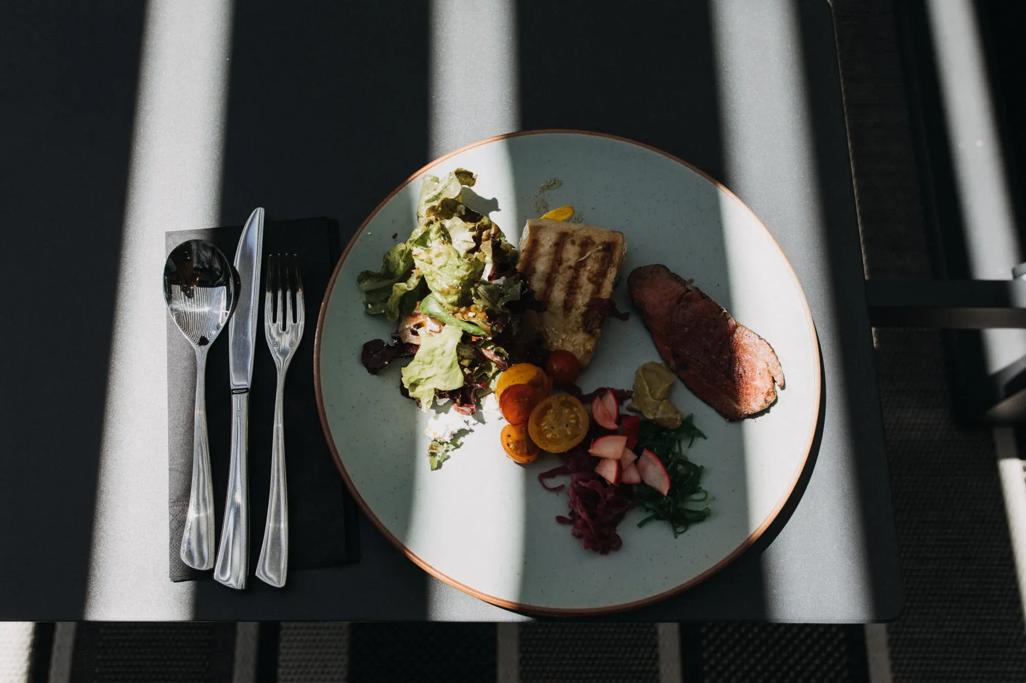 Plate of grilled meat, salad, cherry tomatoes, pickled vegetables on a dark table.