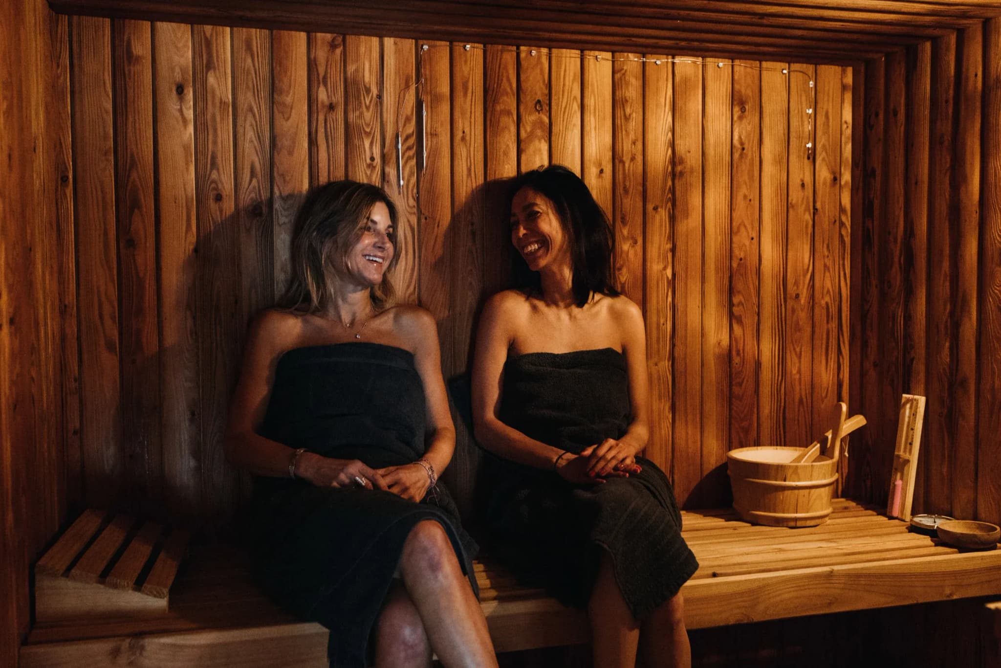 Two guests seated in a wooden sauna, wrapped in dark towels, with a wooden bucket and ladle nearby.
