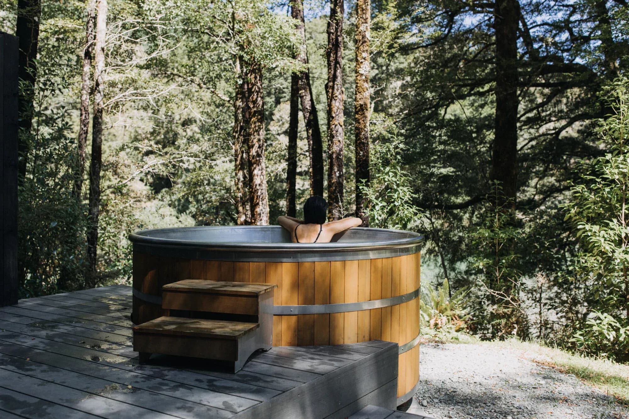 Guest relaxing in a wooden tub surrounded by trees, with water reflecting the natural light.