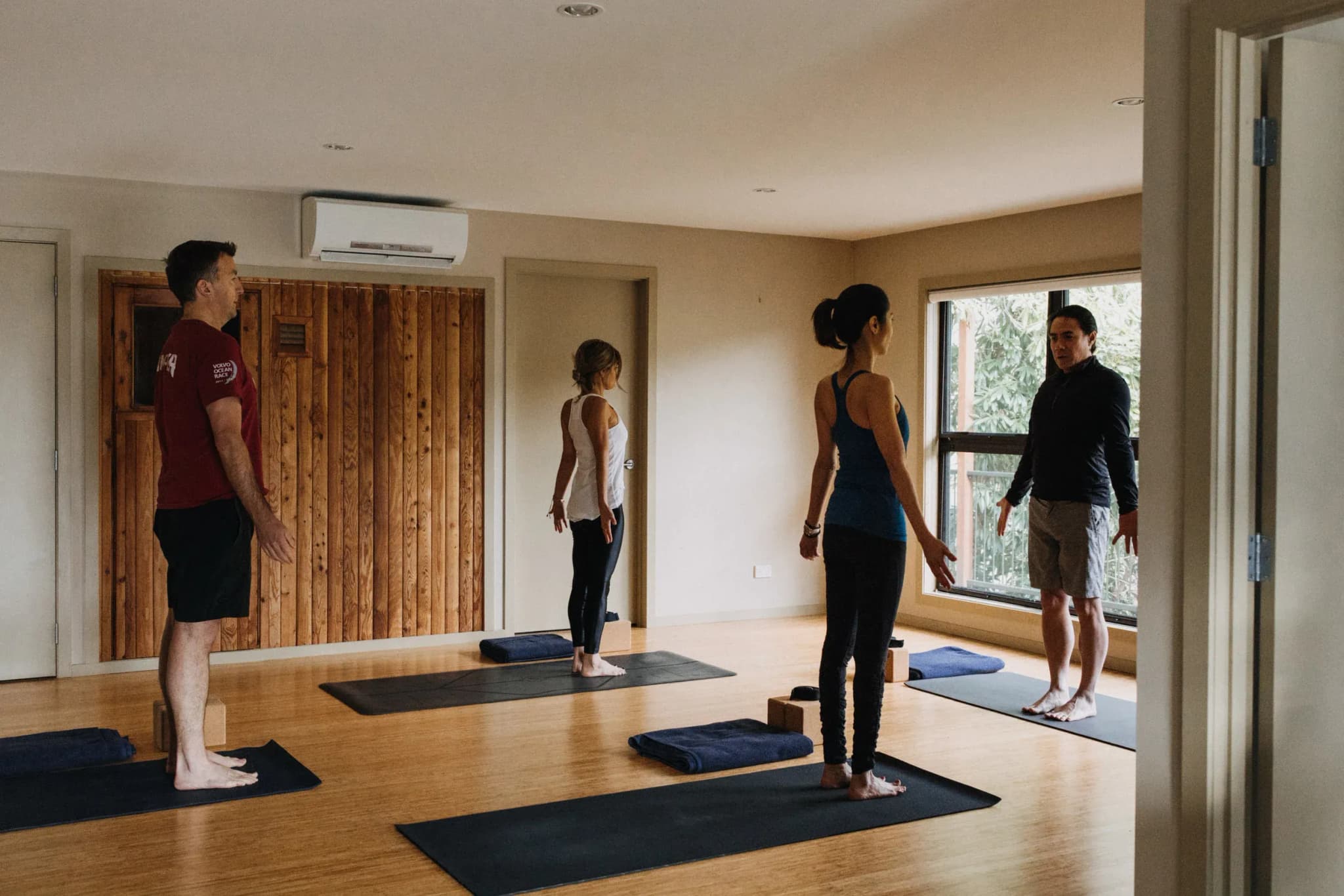 Guests practicing yoga in a wooden-floored studio with mats, blocks, and large windows overlooking greenery.