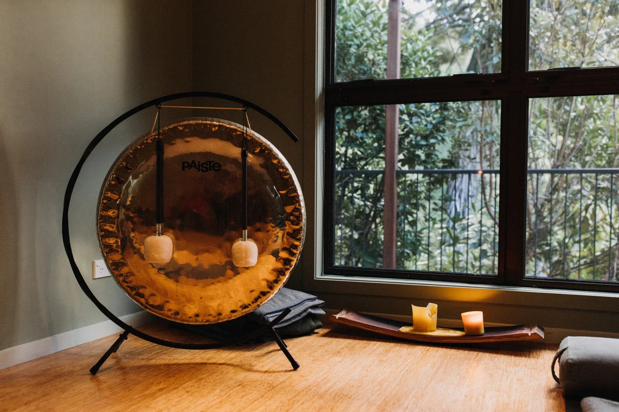 Gong with soft mallets on a stand, beside a window with greenery outside and candles on a wooden tray.