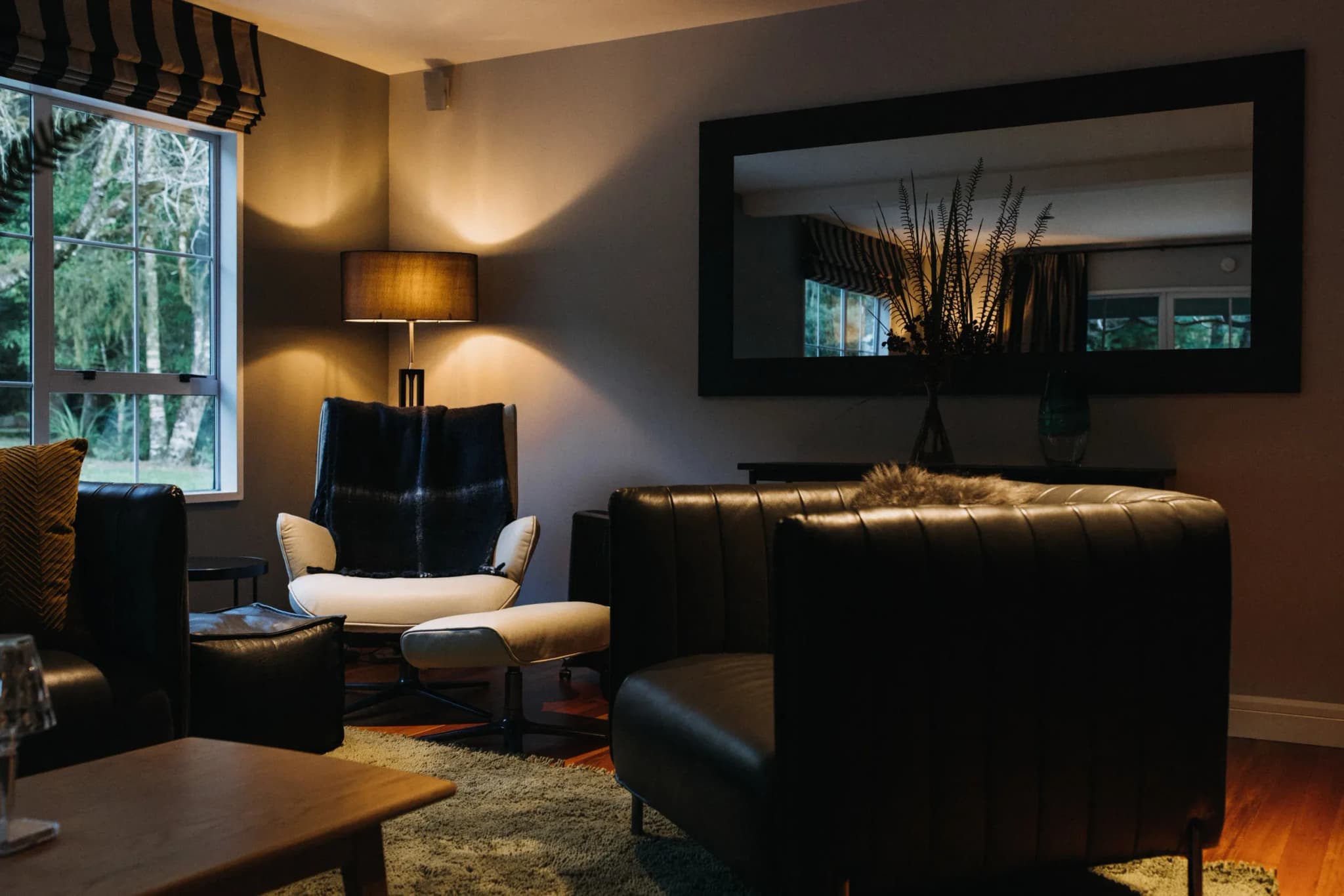 Seating area with black leather armchairs, a white accent chair, a wooden coffee table, and a floor lamp, framed by a large mirror.