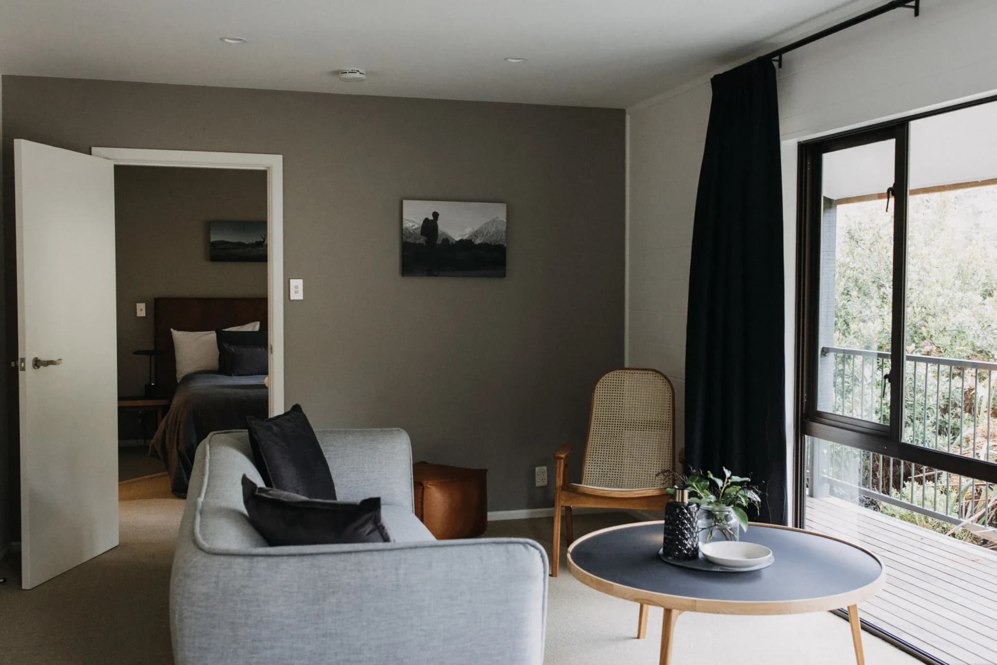 Living area with a grey sofa, wooden chair, coffee table, and an open door leading to a bedroom.