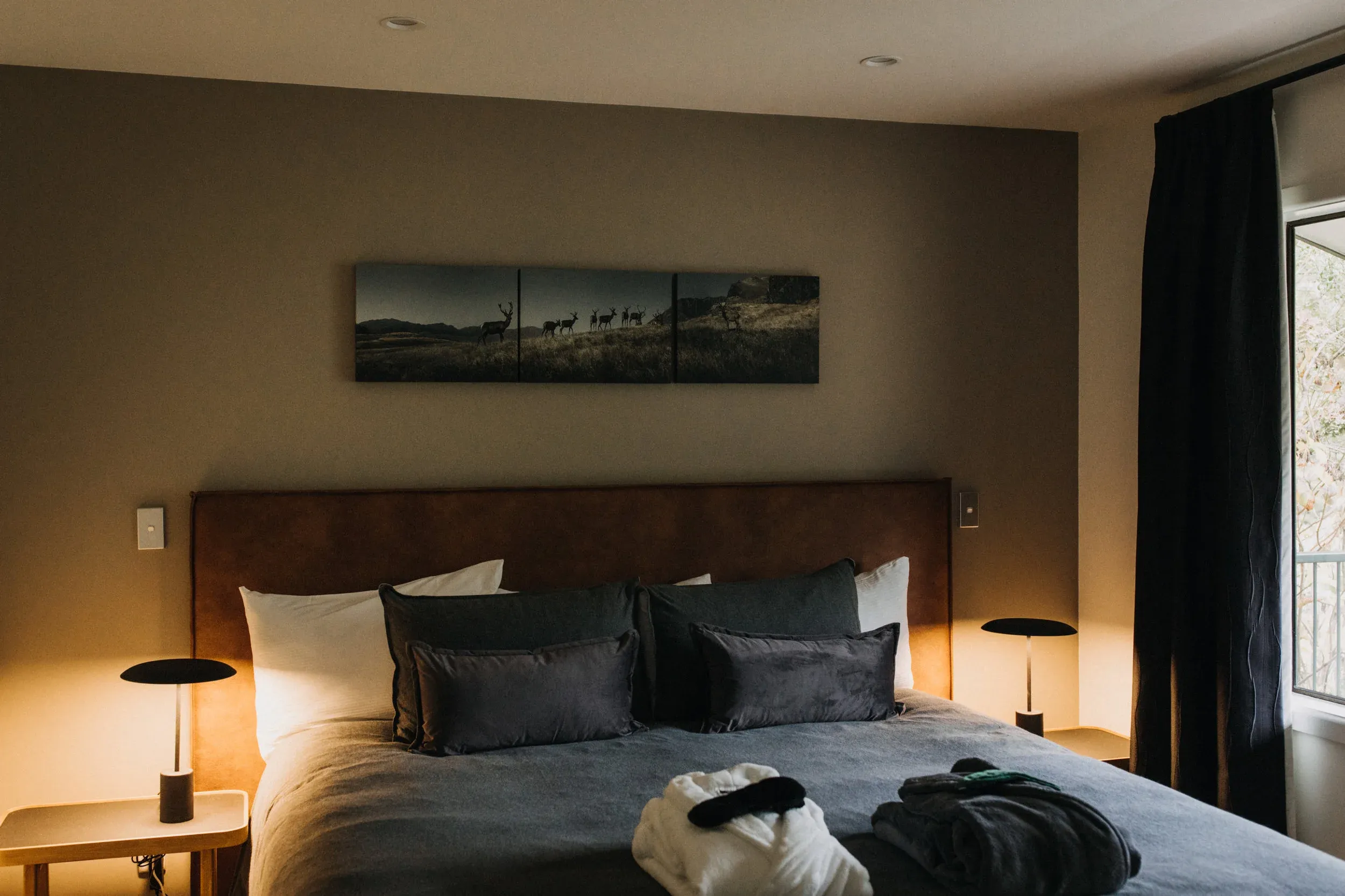 King-sized bed with dark grey and white bedding, flanked by bedside lamps, and a landscape artwork above.