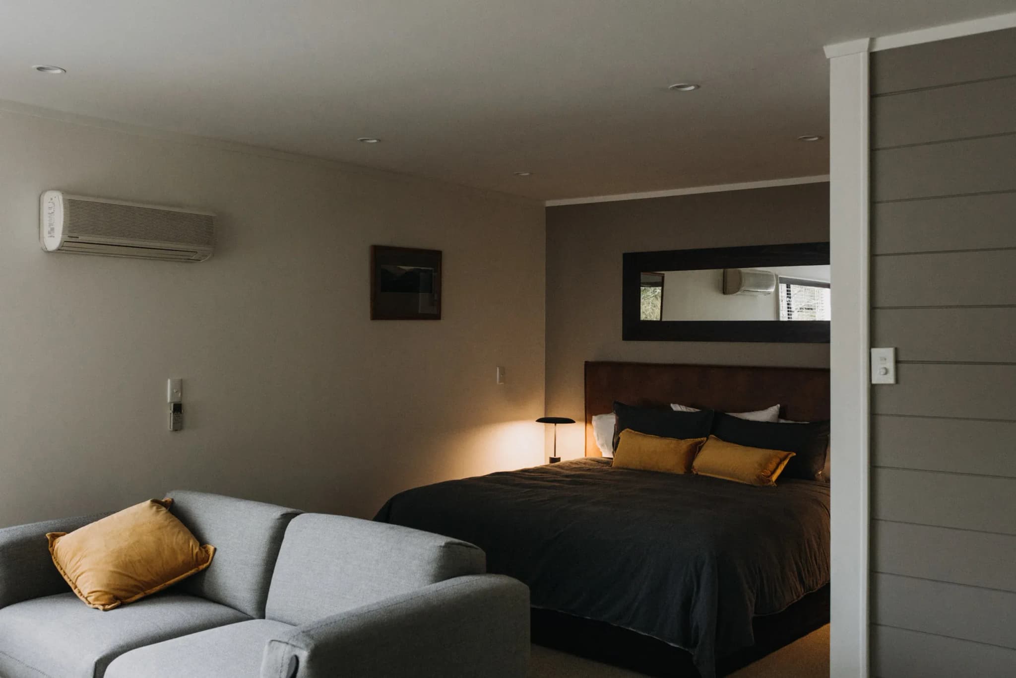 Self-contained room with a grey sofa, bed with dark bedding, yellow cushions, and warm lighting.