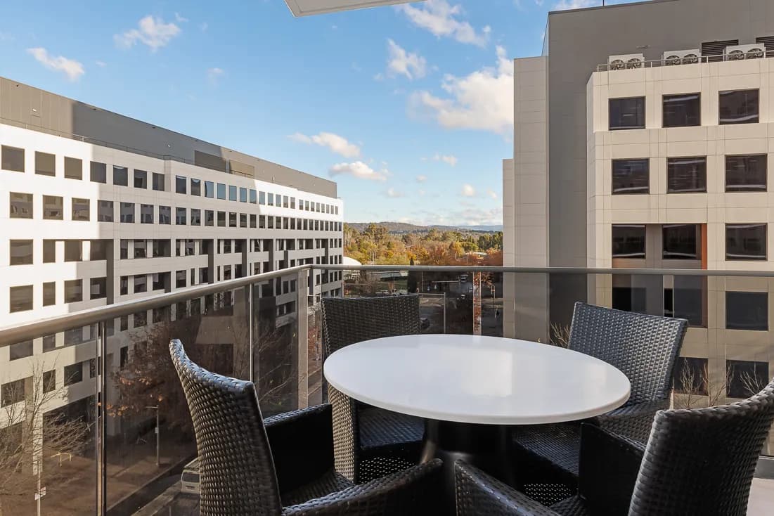 Balcony seating area with a round white table and black wicker chairs, overlooking urban buildings and distant hills under a blue sky.