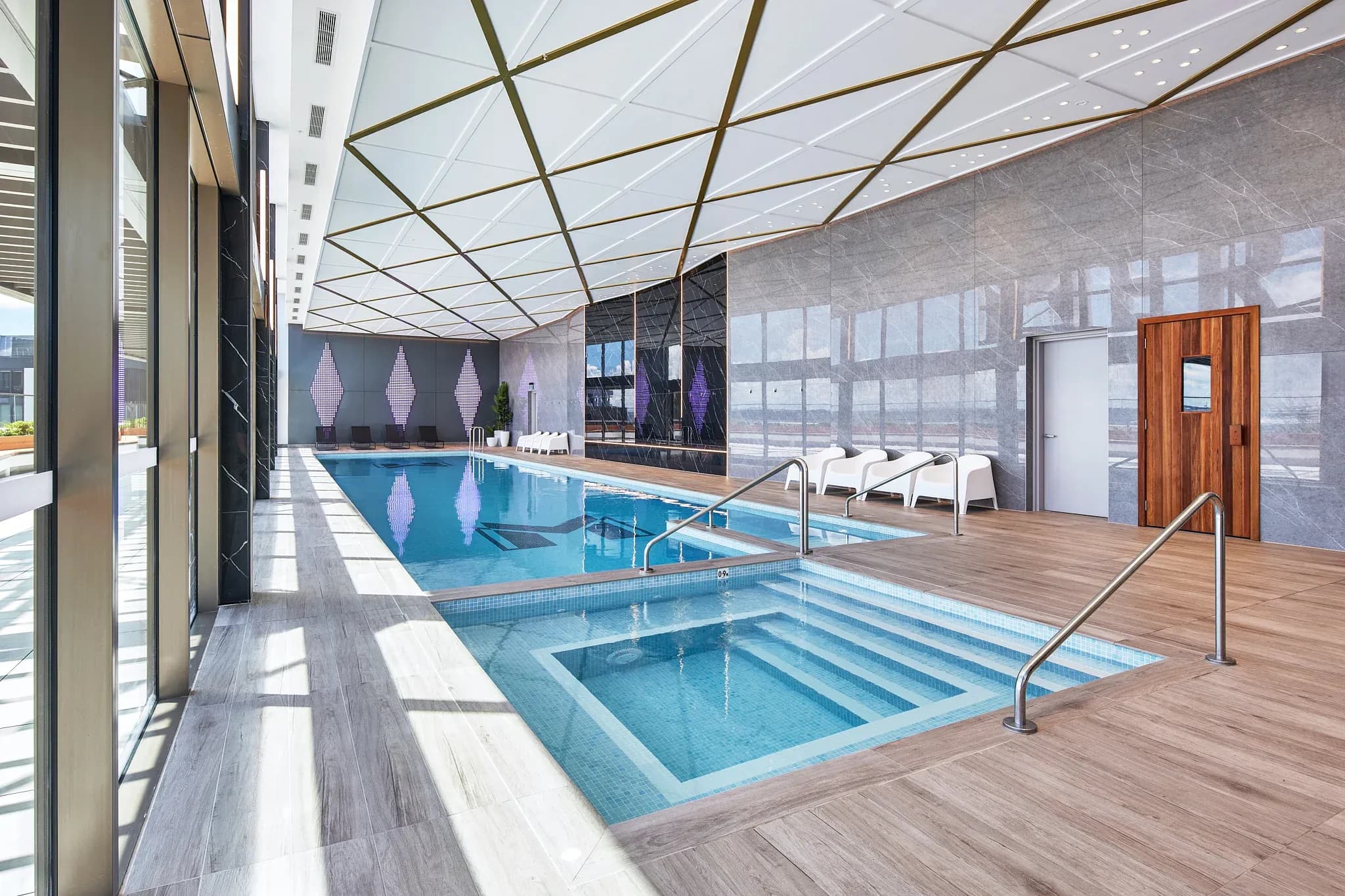 Indoor heated pool area with tiled flooring, lounge chairs, and large windows under a geometric ceiling.