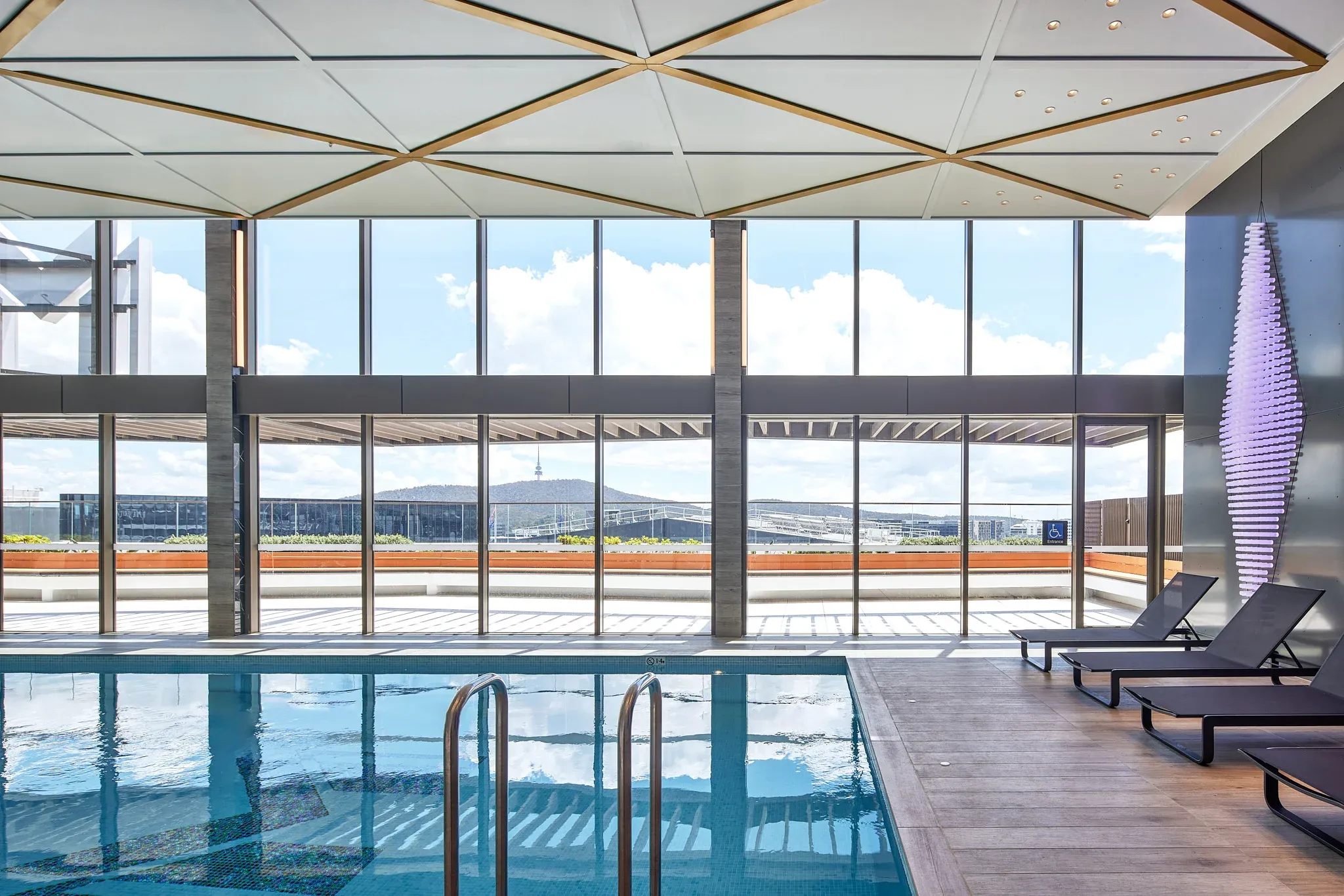 Indoor heated pool with glass walls, lounge chairs, and views of the city skyline and distant hills.