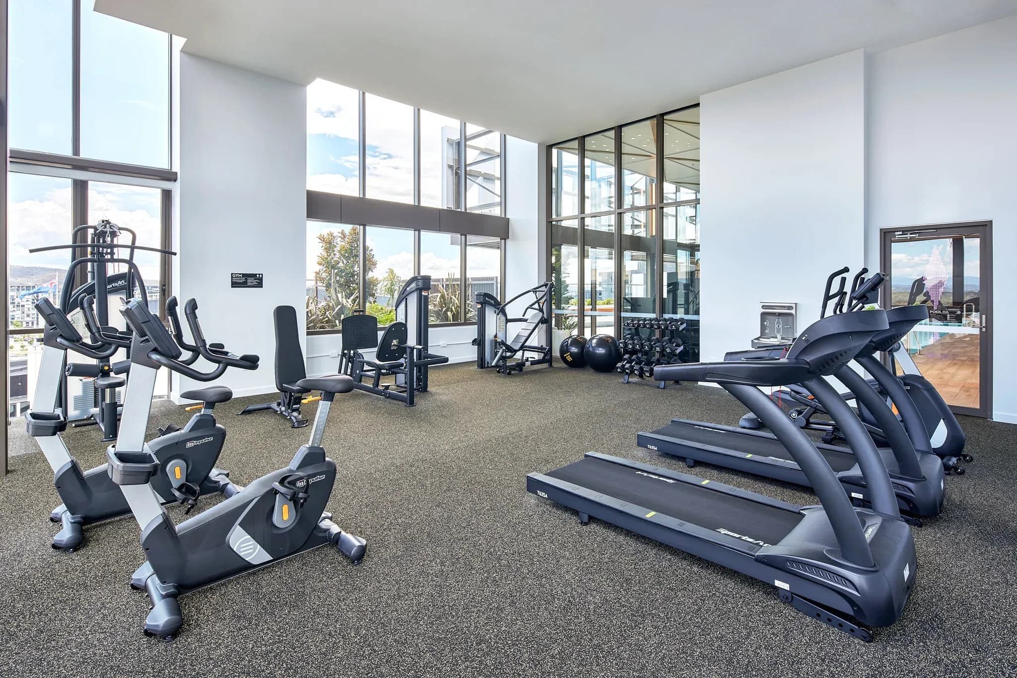 Gym area with exercise bikes, treadmills, weight machines and free weights, featuring large windows and carpeted flooring.