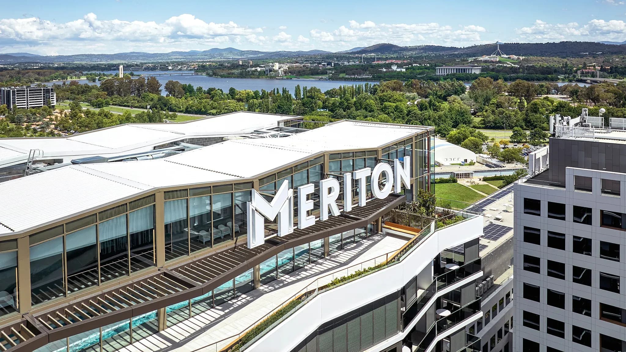 Meriton Suites Canberra signage on a rooftop terrace, overlooking green spaces and a body of water in the distance.