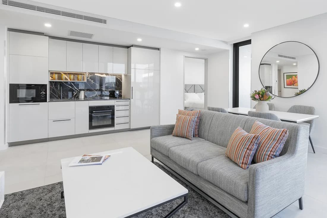 Open-plan suite interior featuring a kitchen with marble backsplash, a grey sofa with striped cushions, and a dining table.