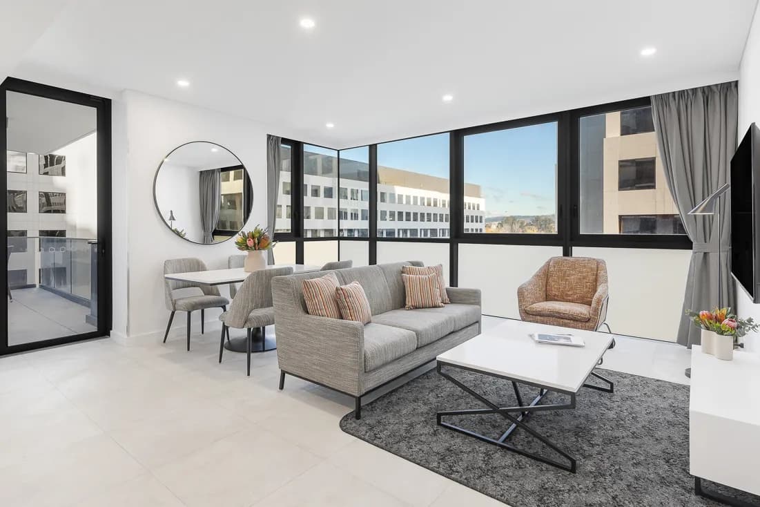 Living area featuring a grey sofa, armchair, dining table, and large windows with city views.