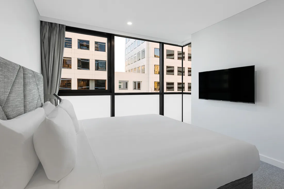 Bedroom with a made bed, gray headboard, televsion and large windows overlooking a neighboring building and cityscape.