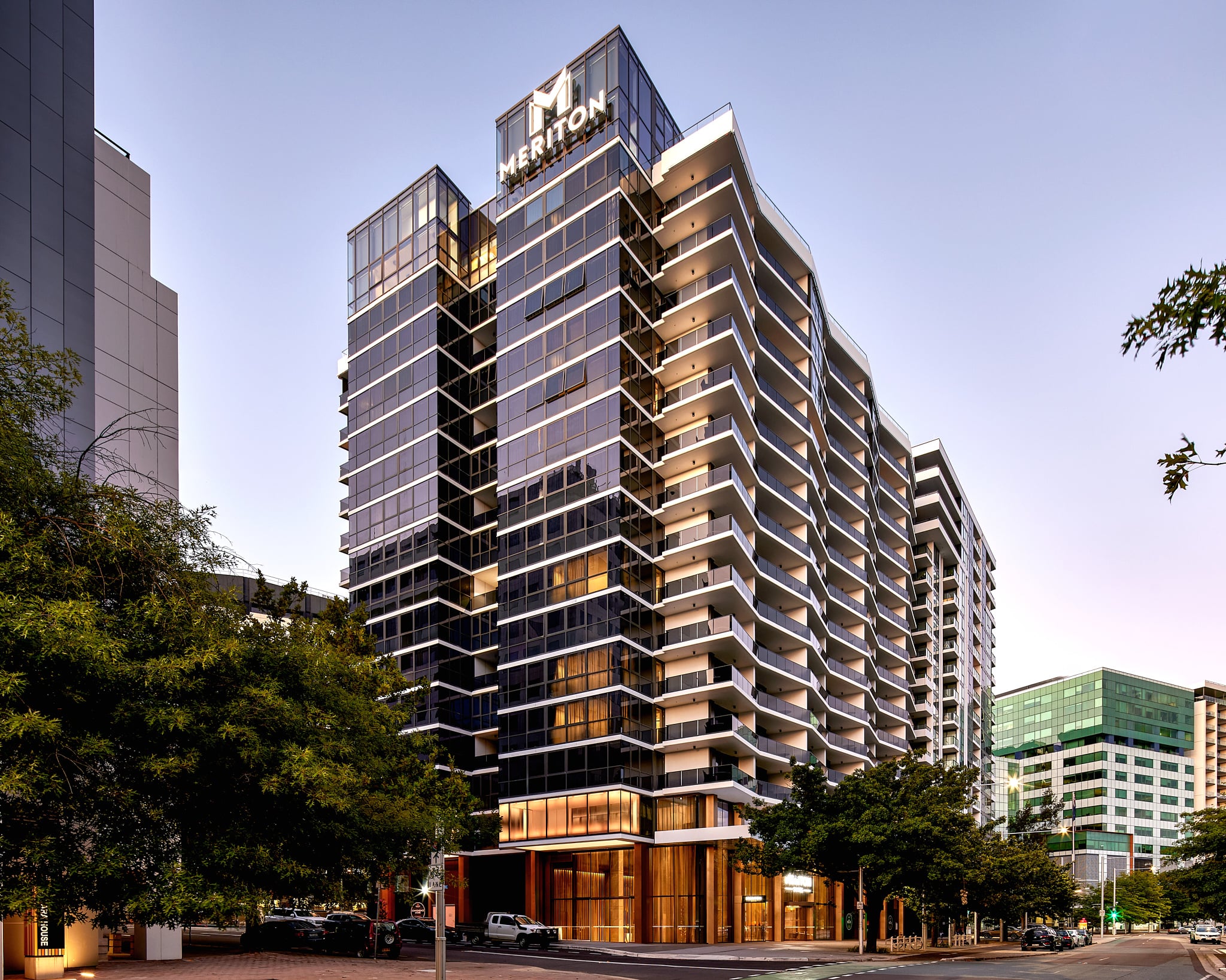 Meriton Suites Canberra exterior featuring a modern glass facade, balconies, and surrounding trees on a city street.