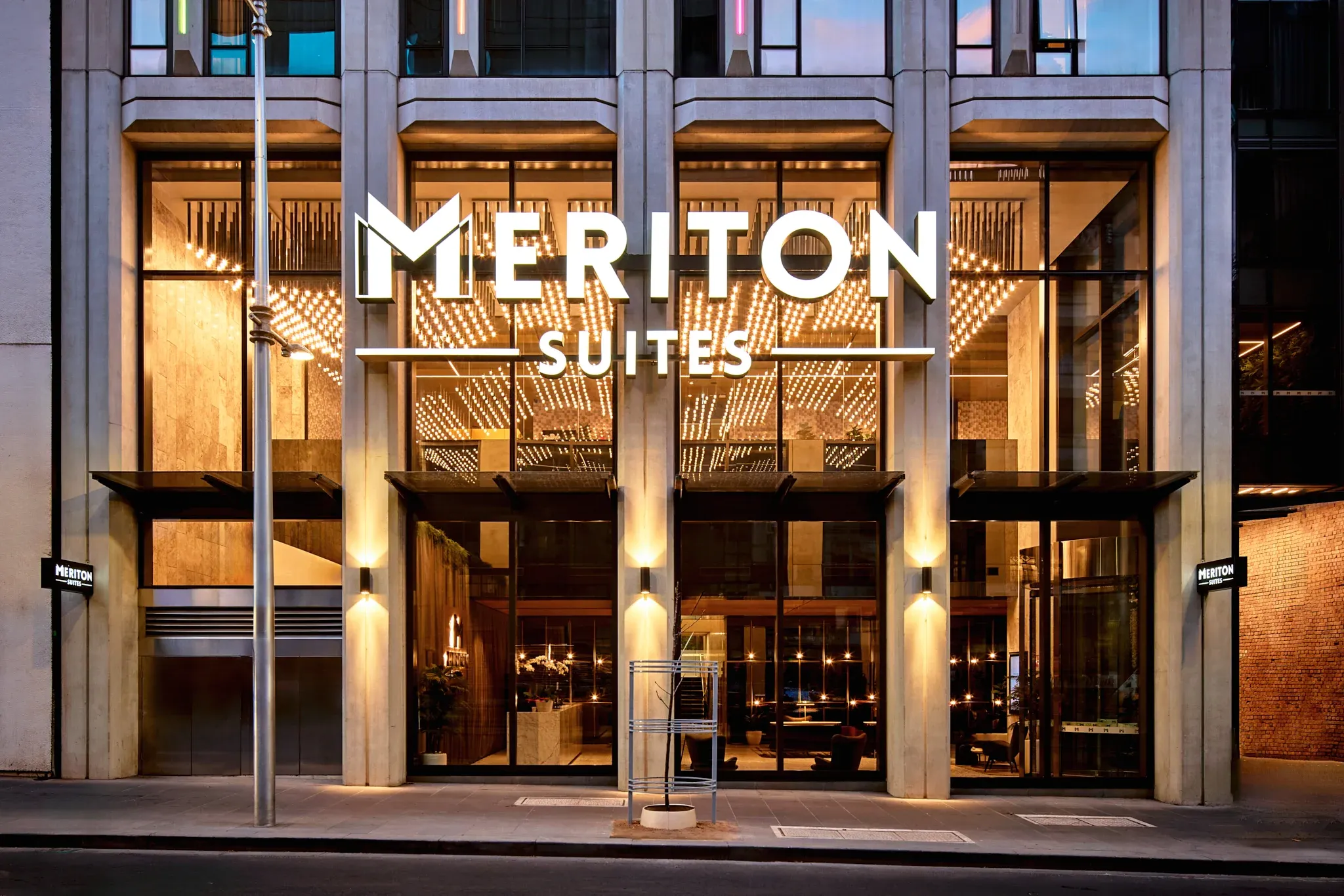 Meriton Suites Melbourne entrance with large glass doors, illuminated signage, and warm lighting reflecting off stone surfaces.