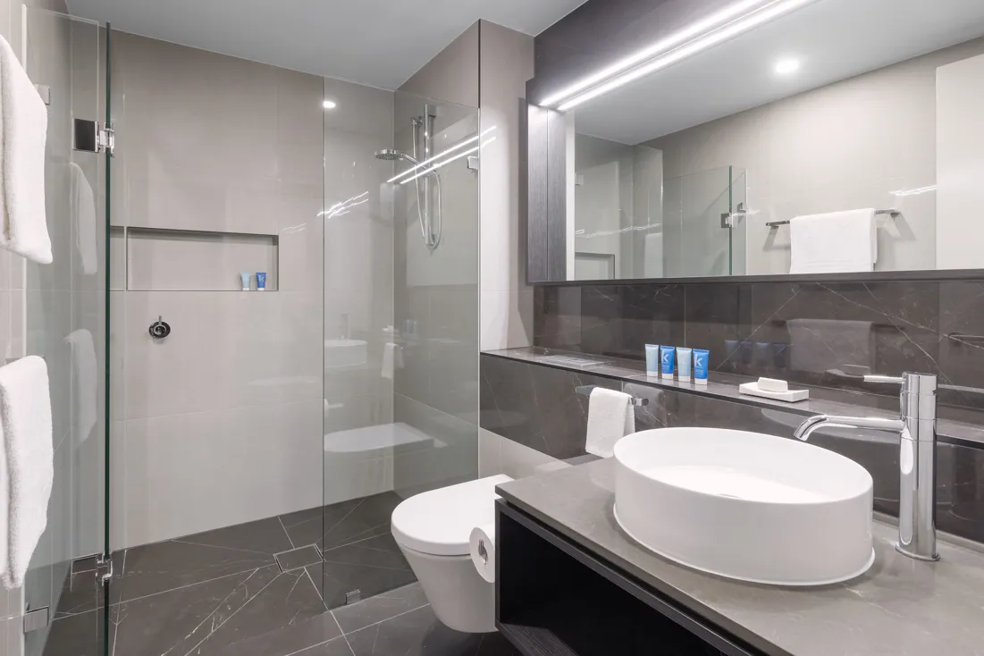 Bathroom interior featuring a glass shower, round basin, dark stone surfaces, and white towels on the wall.